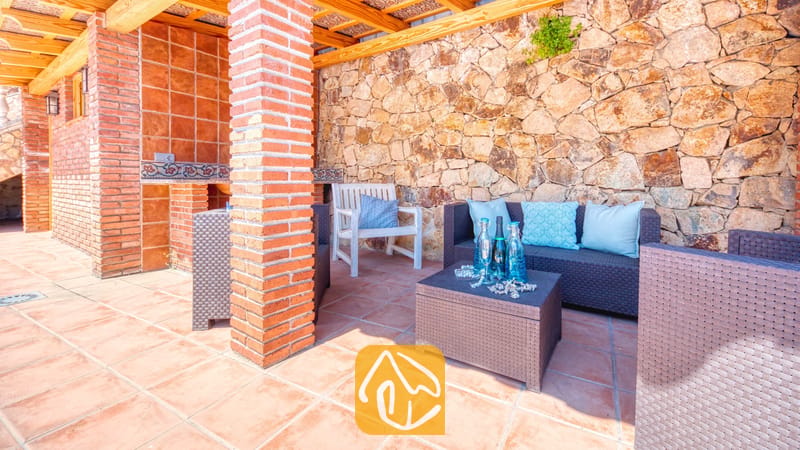 Vakantievilla's Costa Brava Spanje - Villa Dolce Vita - Overdekt buitenterras bij Villa Dolce Vita met bruine rieten zitmeubels, blauwe kussens, een salontafel met glazen flessen en een rustieke stenen muur.