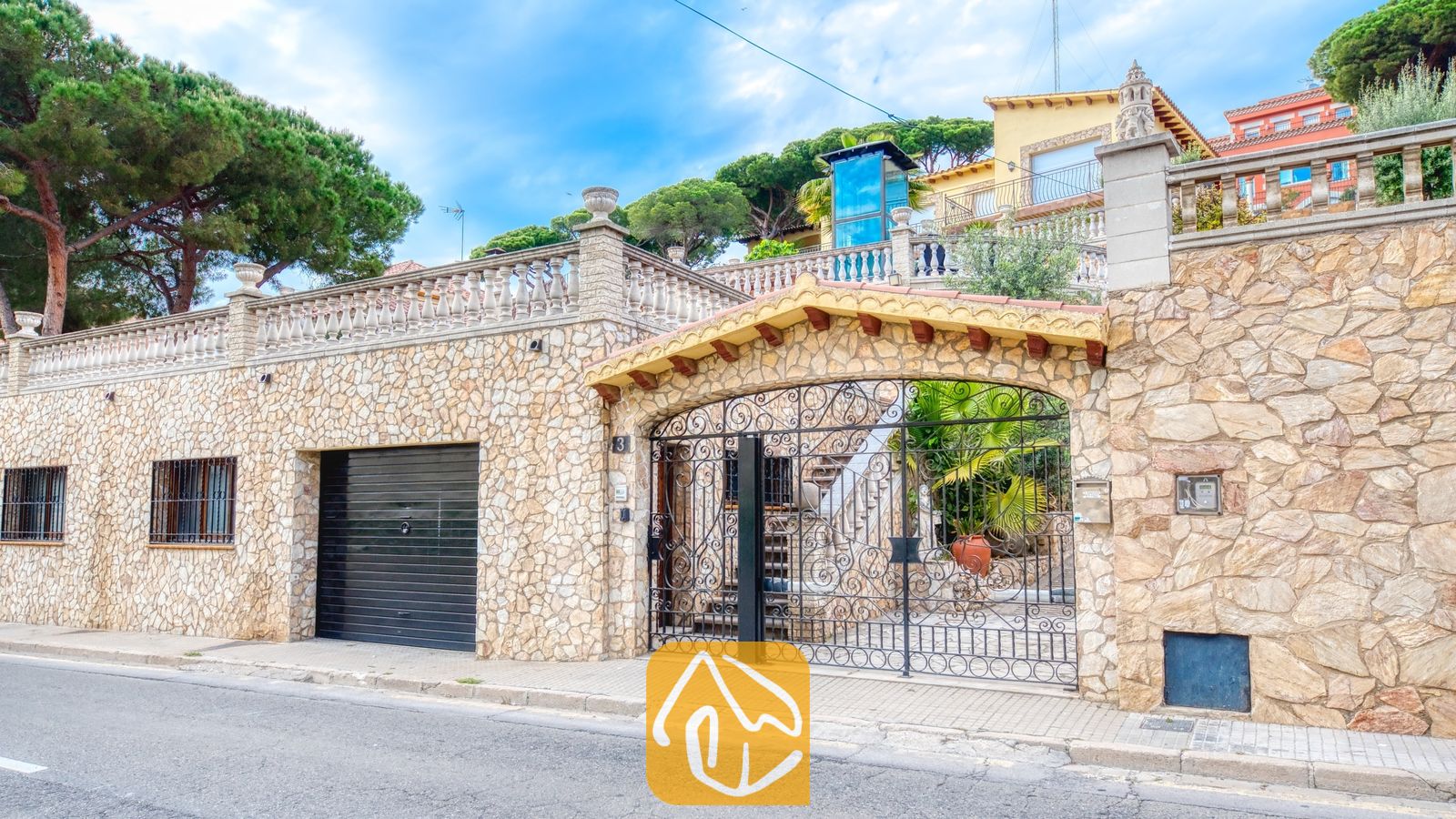 Vakantievilla's Costa Brava Spanje - Villa Dolce Vita - Natuurstenen buitenmuur van Villa Dolce Vita met een zwart smeedijzeren toegangspoort, een bijpassende garagedeur en een terras met stenen balustrades.