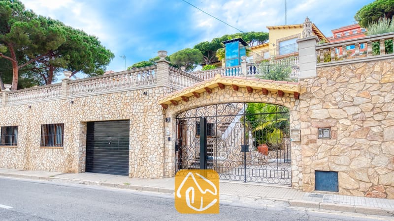 Villas de vacances Costa Brava Espagne - Villa Dolce Vita - Mur extérieur en pierre de la Villa Dolce Vita mettant en valeur un portail d'entrée en fer forgé noir, une porte de garage assortie et une terrasse dotée de balustrades en pierre.