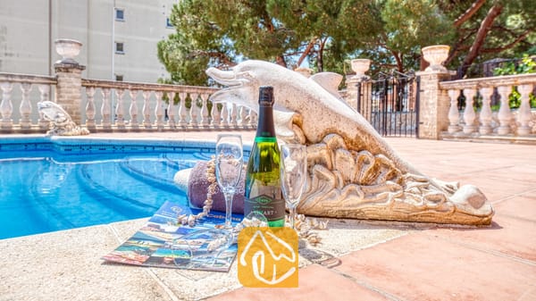 Vakantievilla's Costa Brava Spanje - Villa Dolce Vita - Fles champagne en twee glazen op een betegeld zwembadterras naast een stenen dolfijnenstandbeeld bij Villa Dolce Vita