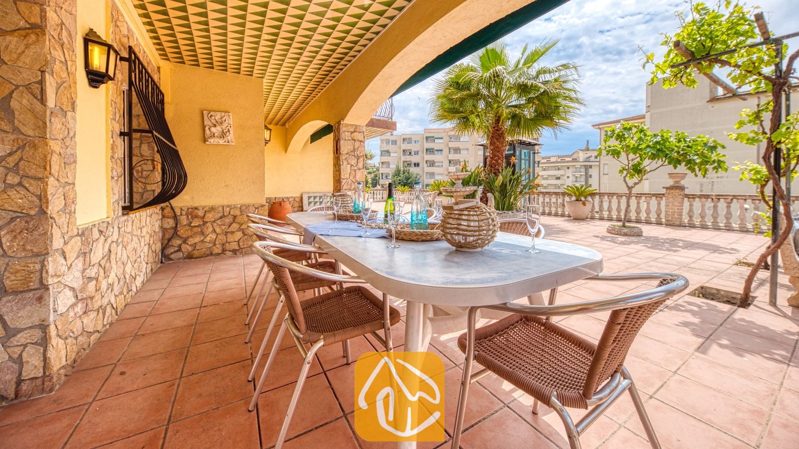 Vakantievilla's Costa Brava Spanje - Villa Dolce Vita - Overdekt terras bij Villa Dolce Vita met een lange eettafel met gevlochten stoelen, stenen muren en een gedecoreerd plafond onder een zonnige hemel.