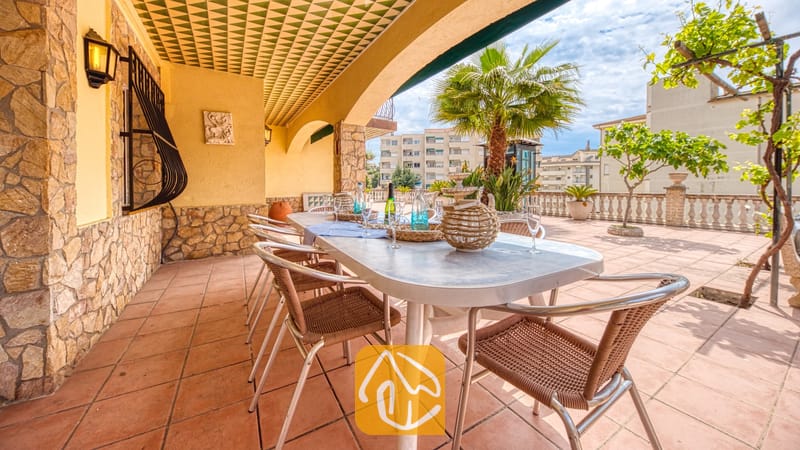 Vakantievilla's Costa Brava Spanje - Villa Dolce Vita - Overdekt terras bij Villa Dolce Vita met een lange eettafel met gevlochten stoelen, stenen muren en een gedecoreerd plafond onder een zonnige hemel.