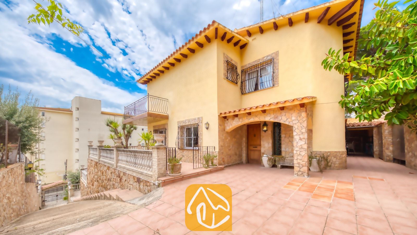 Vakantievilla's Costa Brava Spanje - Villa Dolce Vita - Buitenaanzicht van Villa Dolce Vita met een gele gevel, een pannendak, stenen veranda-ondersteuning en een verhard terras onder een blauwe lucht.