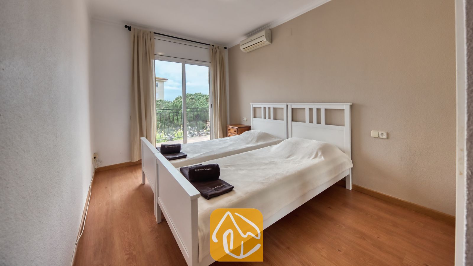Vakantievilla's Costa Brava Spanje - Villa Dolce Vita - Slaapkamer met twee witte houten eenpersoonsbedden, houten vloeren en een balkondeur met uitzicht op bomen.