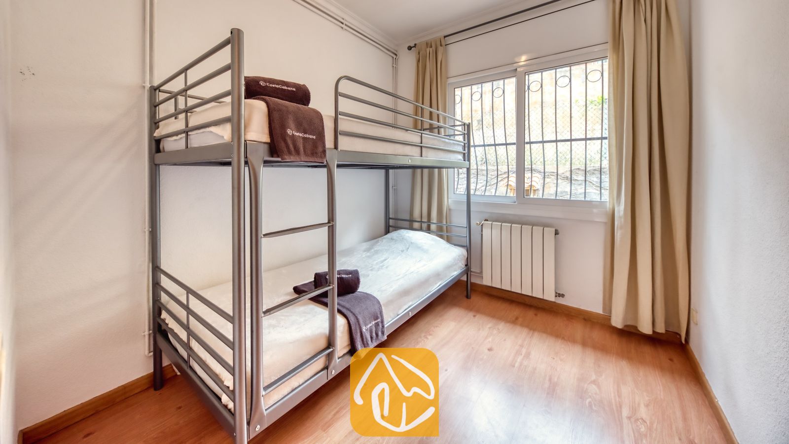 Vakantievilla's Costa Brava Spanje - Villa Dolce Vita - Slaapkamer met een grijs metalen stapelbed met witte matrassen en opgevouwen bruine handdoeken, tegen een witte muur met een raam en beige gordijnen.