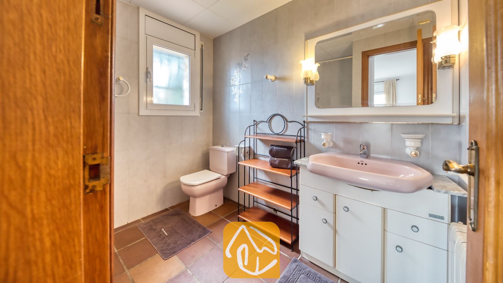 Vakantievilla's Costa Brava Spanje - Villa Dolce Vita - Badkamer met een roze wastafelmeubel, bijpassend toilet, betegelde muren en een metalen wandrek met houten planken