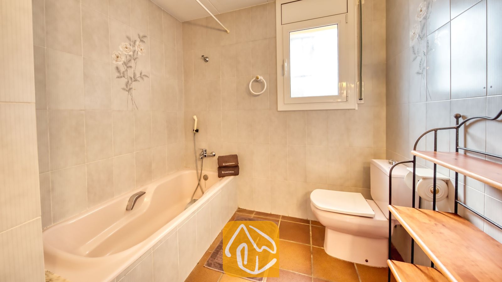 Vakantievilla's Costa Brava Spanje - Villa Dolce Vita - Badkamer met een beige ligbad, een wit toilet, betegelde muren met bloemmotieven en een houten opbergrek met meerdere niveaus en een metalen frame.