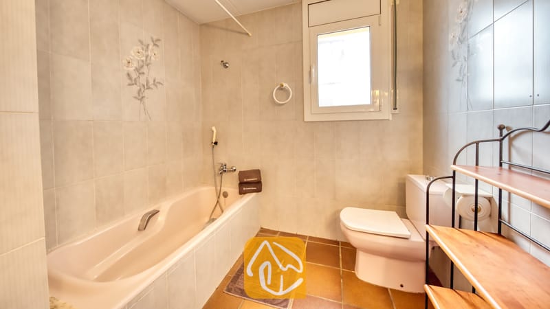 Villas de vacances Costa Brava Espagne - Villa Dolce Vita - Salle de bains comprenant une baignoire beige, des toilettes blanches, des murs carrelés ornés de motifs floraux et une étagère en bois à plusieurs niveaux dotée d'une structure en métal.