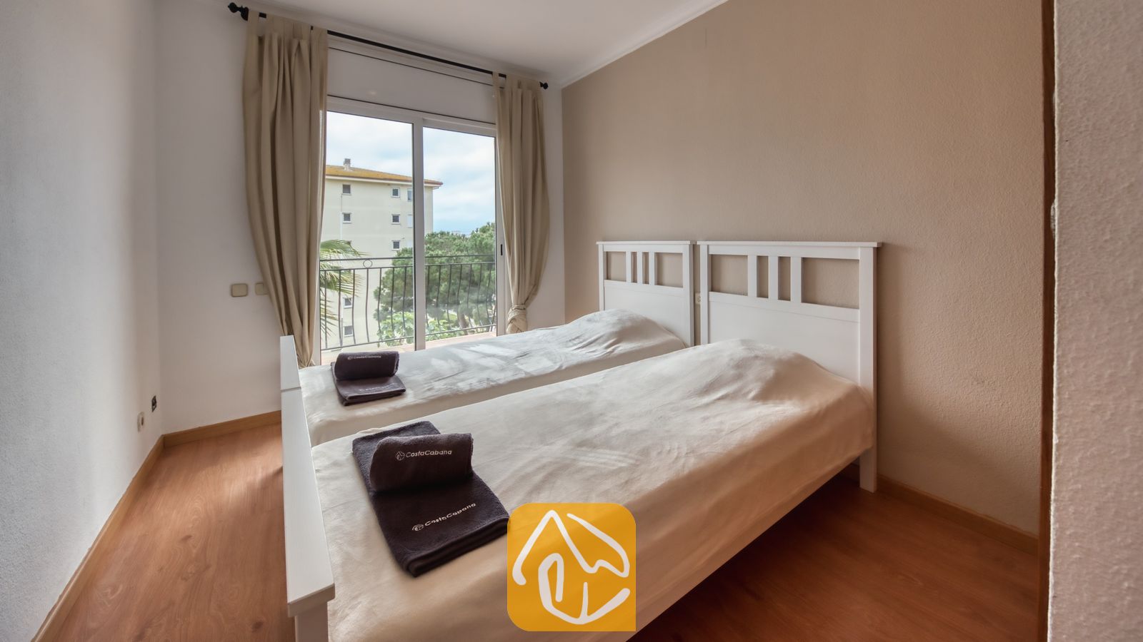 Vakantievilla's Costa Brava Spanje - Villa Dolce Vita - Slaapkamer met twee witte, houten eenpersoonsbedden, beige gordijnen en glazen schuifpui naar een balkon met uitzicht op bomen.