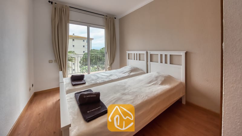 Villas de vacances Costa Brava Espagne - Villa Dolce Vita - Chambre à coucher avec deux lits simples à cadre en bois blanc, rideaux beiges et portes coulissantes en verre s'ouvrant sur un balcon avec vue sur les arbres.