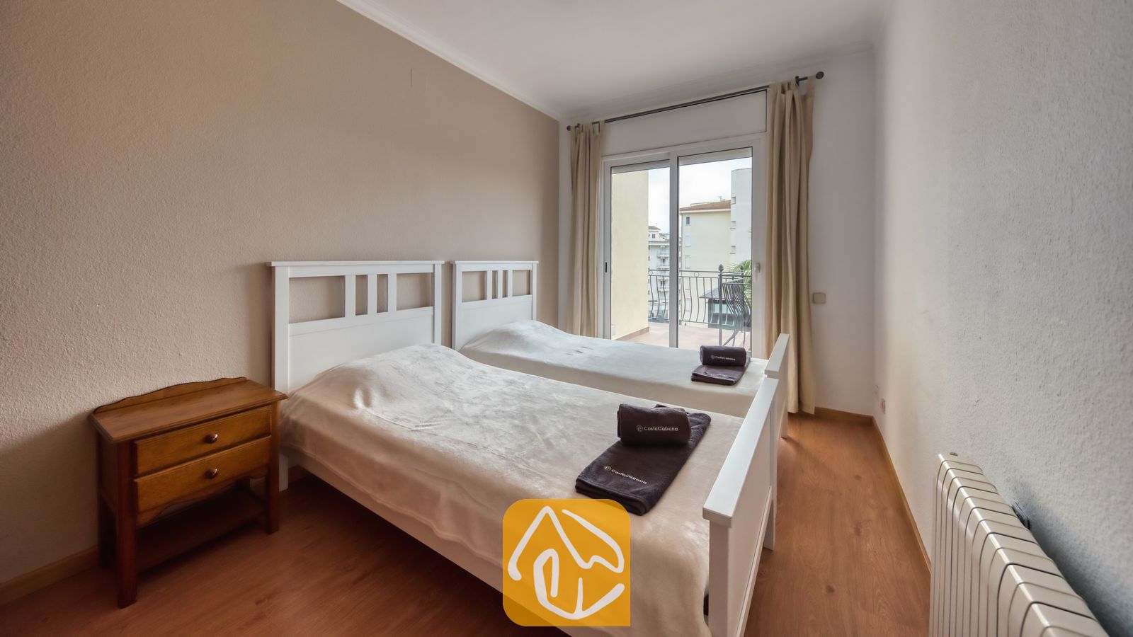 Vakantievilla's Costa Brava Spanje - Villa Dolce Vita - Slaapkamer met twee witte eenpersoonsbedden, een houten nachtkastje en een schuifpui die toegang biedt tot een balkon.