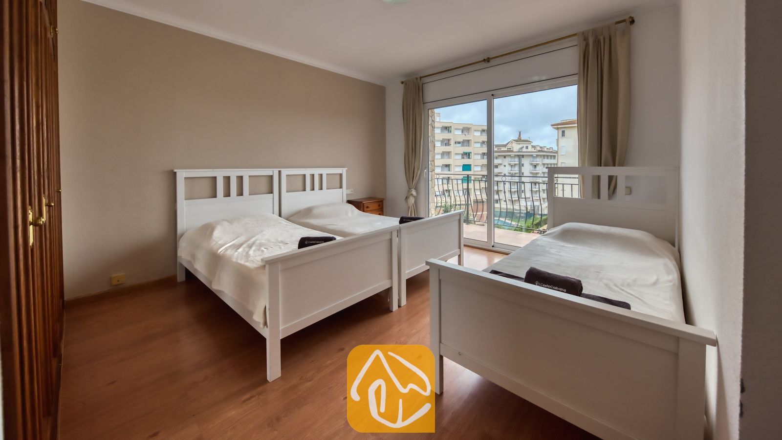 Vakantievilla's Costa Brava Spanje - Villa Dolce Vita - Slaapkamer met drie witte eenpersoonsbedden, houten vloeren en een glazen schuifpui die uitkomt op een balkon met uitzicht op de stad.