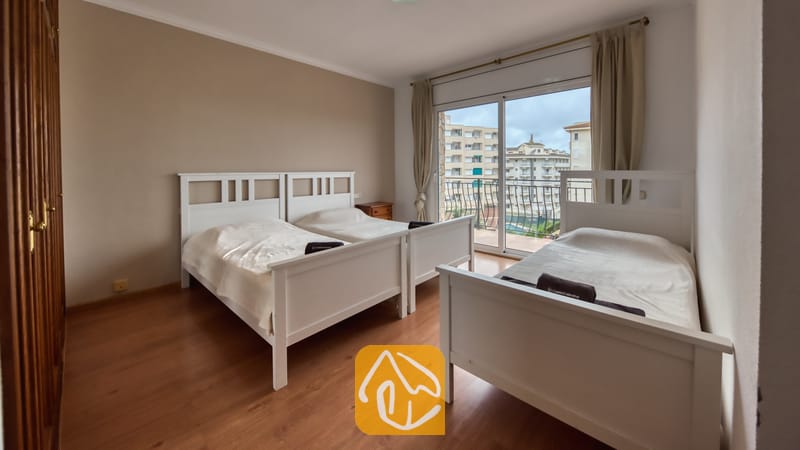 Villas de vacances Costa Brava Espagne - Villa Dolce Vita - Chambre avec trois lits simples blancs, parquets en bois et porte-fenêtre coulissante s'ouvrant sur un balcon avec vue sur la ville.