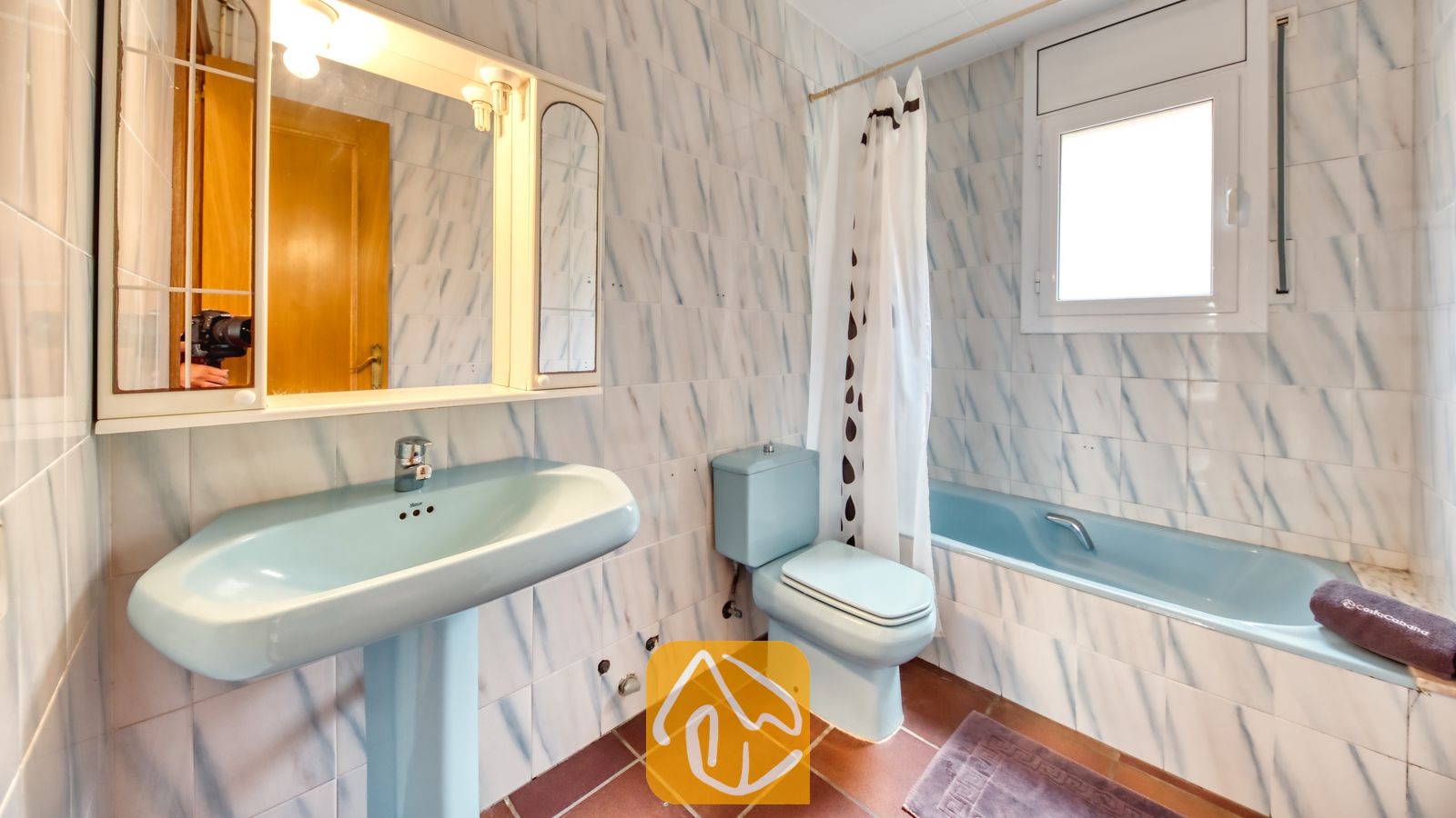 Vakantievilla's Costa Brava Spanje - Villa Dolce Vita - Badkamer met een lichtblauwe zuilwastafel, bijpassend toilet en ligbad met een wit douchegordijn met patroon, en een wandspiegel met ingebouwde verlichting.