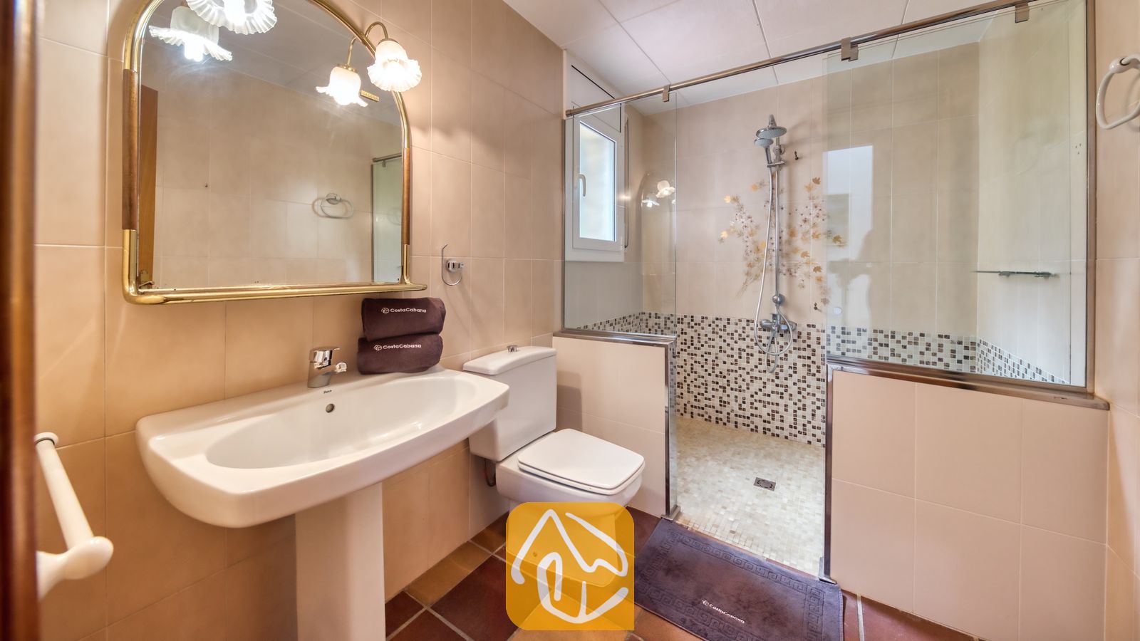 Vakantievilla's Costa Brava Spanje - Villa Dolce Vita - Badkamer met een witte vrijstaande wastafel, toilet en een glazen inloopdouche met mozaïek tegelaccenten en een koperen spiegel aan de beige getegelde muren.