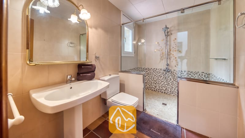 Villas de vacances Costa Brava Espagne - Villa Dolce Vita - Salle de bains comprenant un lavabo sur pied blanc, des toilettes, et une douche à l'italienne vitrée avec des touches de mosaïque et un miroir encadré de laiton sur des murs carrelés de beige.