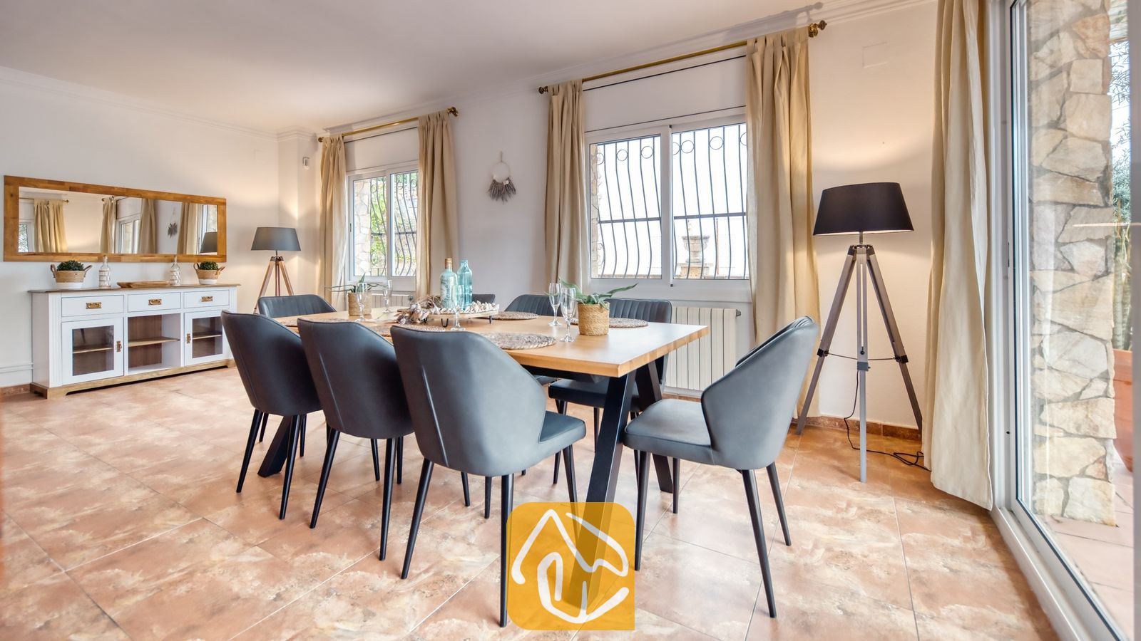 Vakantievilla's Costa Brava Spanje - Villa Dolce Vita - Eetkamer met een houten tafel, donkergrijze gestoffeerde stoelen, tegelvloer, staande lampen met driepoot en ramen met ecrukleurige gordijnen.