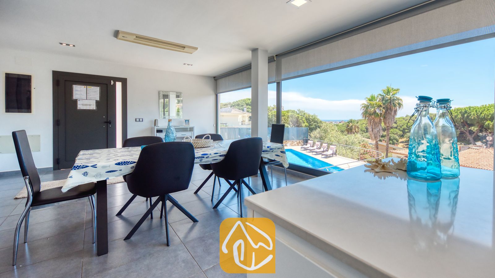 Vakantievilla's Costa Brava Spanje - Villa SummerDream - Eethoek binnen in Villa SummerDream, voorzien van een donkere eettafel met zwarte stoelen, een tafelkleed met patroon en grote kamerhoge ramen met uitzicht op een buitenzwembad, terras en palmbomen.