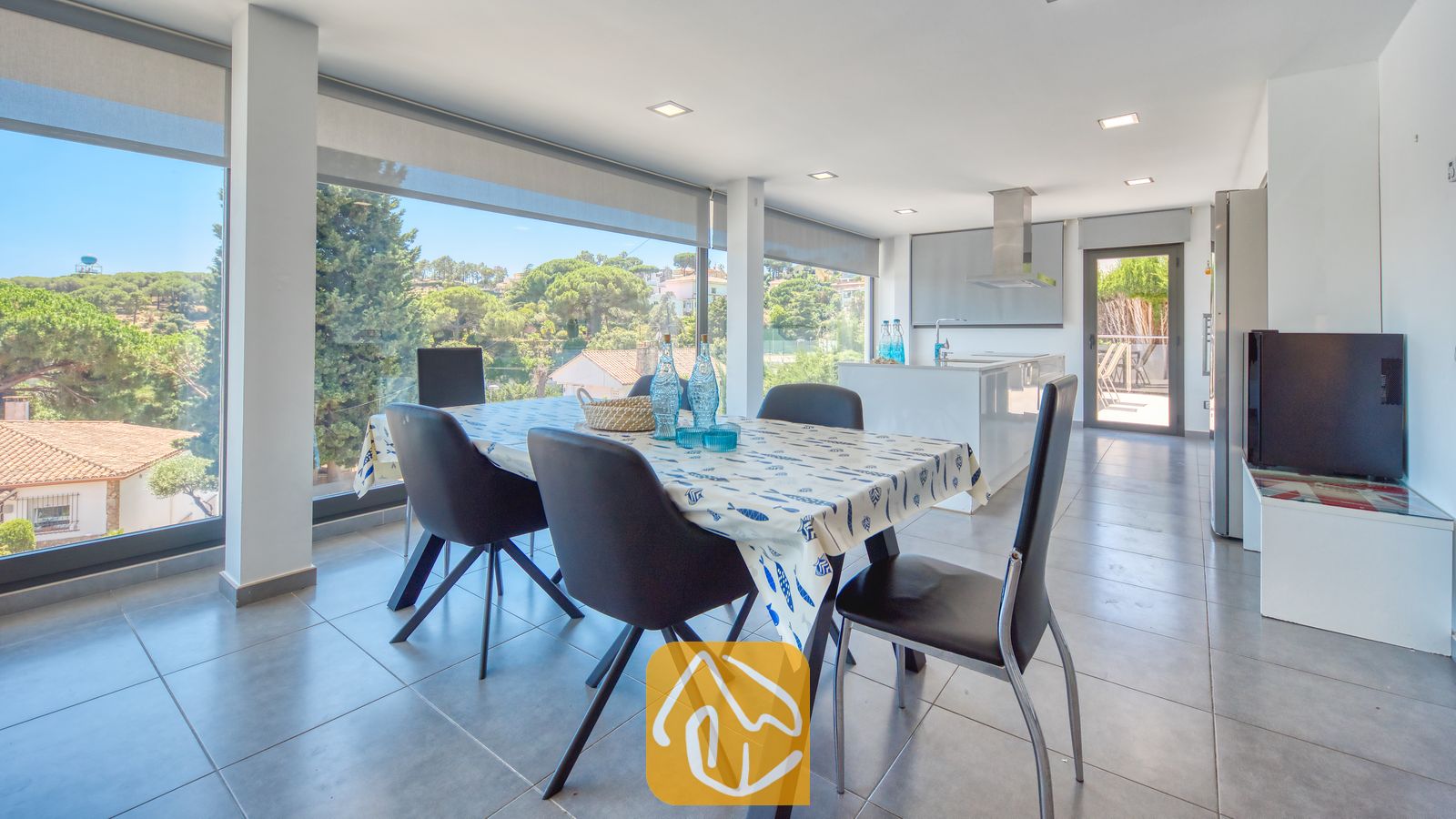 Vakantievilla's Costa Brava Spanje - Villa SummerDream - Eethoek in Villa SummerDream met een tafel en zwarte stoelen, grote ramen met uitzicht op groene bomen en een open keuken op de achtergrond.