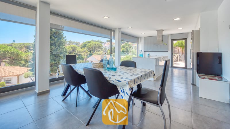 Villas de vacances Costa Brava Espagne - Villa SummerDream - Espace repas à l'intérieur de la Villa SummerDream comprenant une table avec des chaises noires, de grandes baies vitrées offrant une vue sur les arbres verdoyants, et une cuisine ouverte en arrière-plan.
