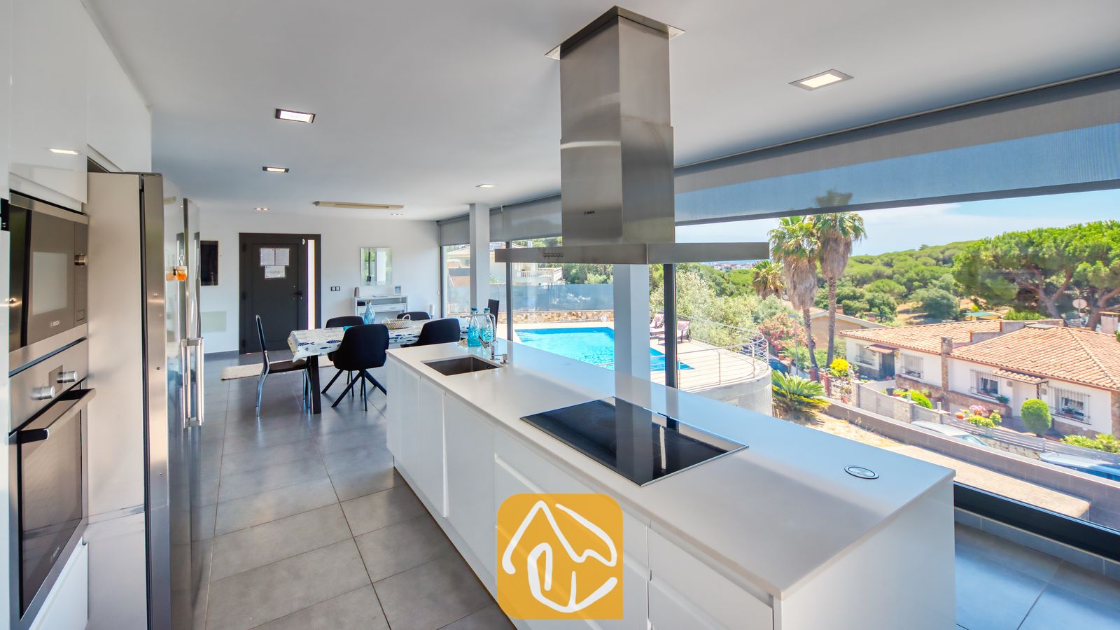 Vakantievilla's Costa Brava Spanje - Villa SummerDream - Moderne keuken met witte aanrechtbladen, een afzuigkap van roestvrij staal en grote ramen met uitzicht op een zwembad en bomen bij Villa SummerDream.