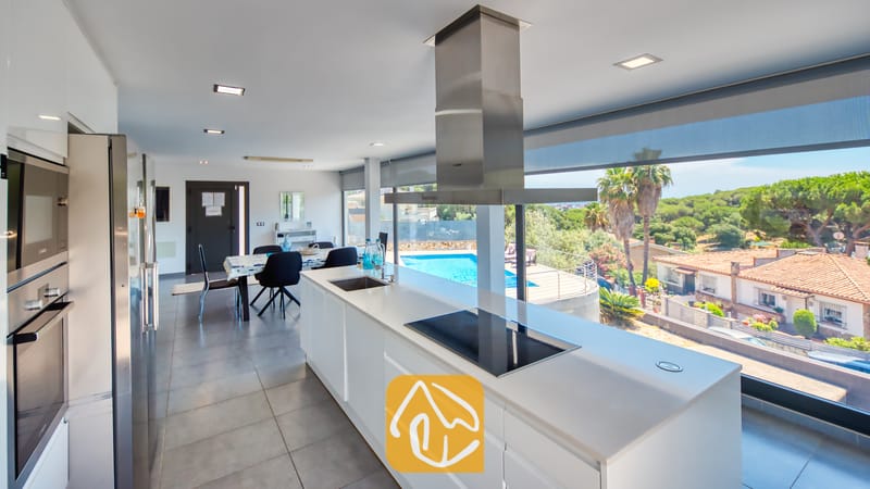 Villas de vacances Costa Brava Espagne - Villa SummerDream - Cuisine moderne dotée de comptoirs blancs, d'une hotte aspirante en acier inoxydable et de grandes fenêtres offrant une vue sur la piscine et les arbres à la Villa SummerDream.