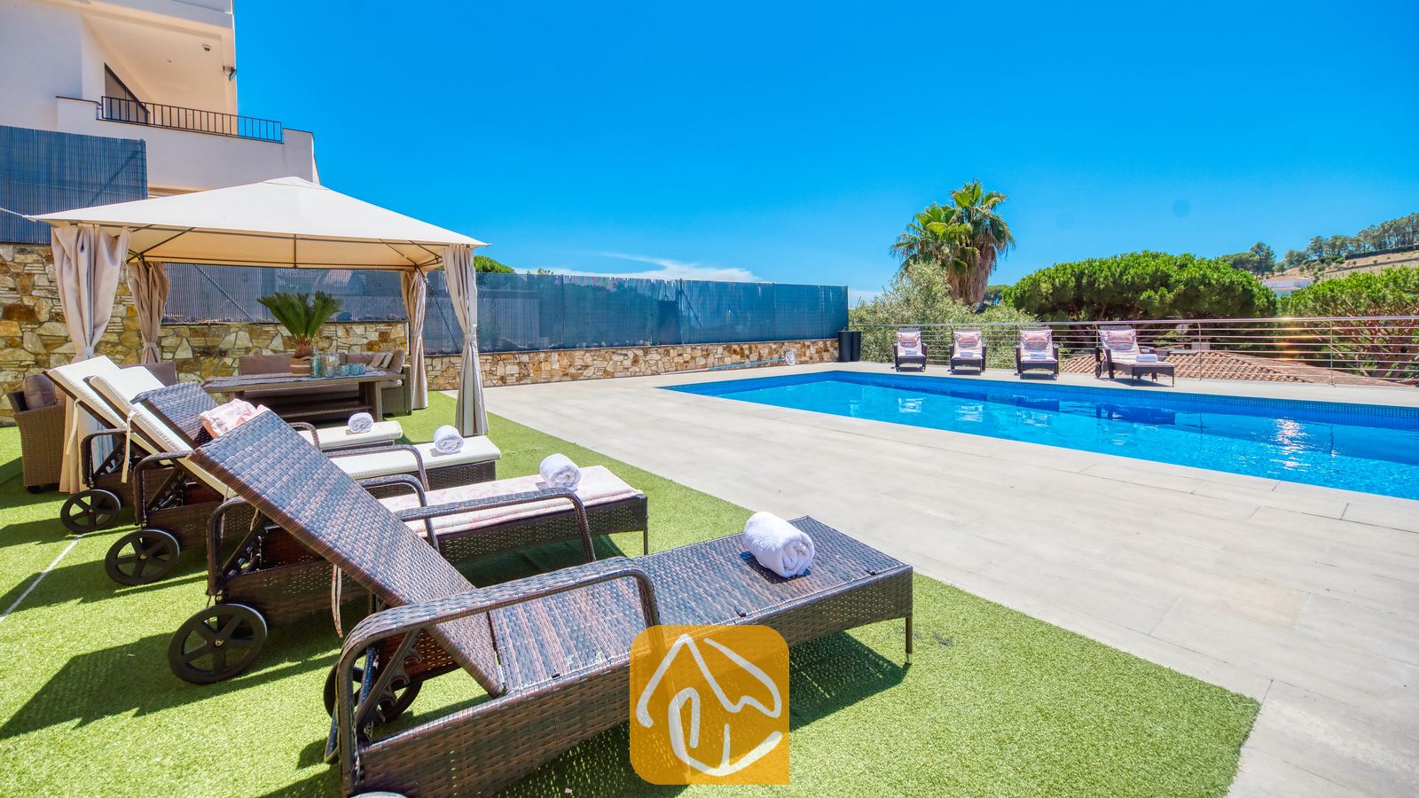 Vakantievilla's Costa Brava Spanje - Villa SummerDream - Het buitenzwembadgedeelte van Villa SummerDream, voorzien van donkerwicker ligbedden met witte handdoeken op kunstgras, een beige prieel en in de verte palmbomen tegen een blauwe hemel.
