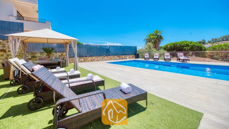 Vakantievilla's Costa Brava Spanje - Villa SummerDream - Het buitenzwembadgedeelte van Villa SummerDream, voorzien van donkerwicker ligbedden met witte handdoeken op kunstgras, een beige prieel en in de verte palmbomen tegen een blauwe hemel.