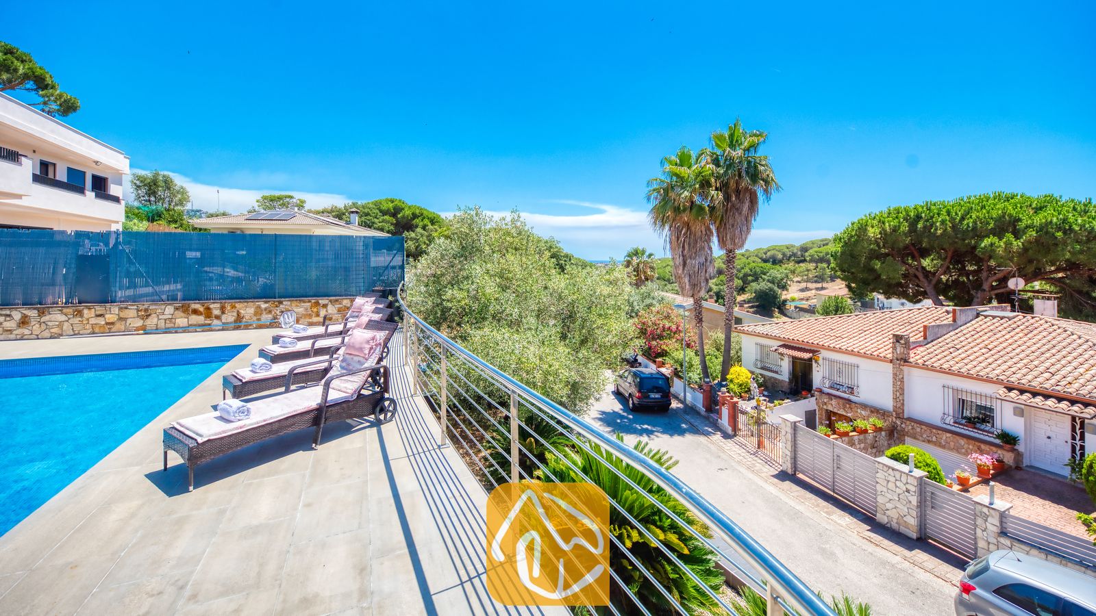 Vakantievilla's Costa Brava Spanje - Villa SummerDream - Zonnig terras bij Villa SummerDream met een rij donkerkleurige wicker ligbedden met kussens en opgerolde handdoeken naast een zwembad, met uitzicht op een straat en palmbomen.