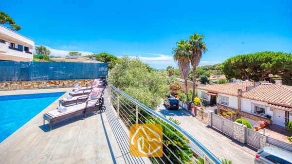 Vakantievilla's Costa Brava Spanje - Villa SummerDream - Zonnig terras bij Villa SummerDream met een rij donkerkleurige wicker ligbedden met kussens en opgerolde handdoeken naast een zwembad, met uitzicht op een straat en palmbomen.