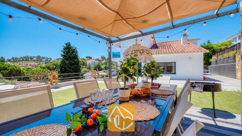 Vakantievilla's Costa Brava Spanje - Villa SummerDream - Eettafel in de buitenlucht onder een beige luifel met sfeerverlichting bij Villa SummerDream, voorzien van een glazen blad, geweven placemats, in pot gekweekte citroenbomen en uitzicht op het landschap.