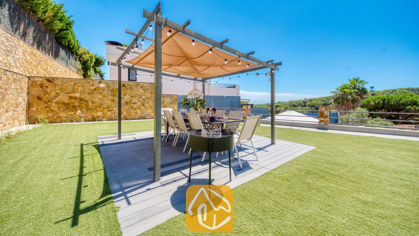 Vakantievilla's Costa Brava Spanje - Villa SummerDream - Eethoek buiten bij Villa SummerDream met een overdekte pergola met sfeerverlichting, een grote eettafel met witte stoelen en een verplaatsbare grill op een houten vlonder, omringd door een groen gazon.