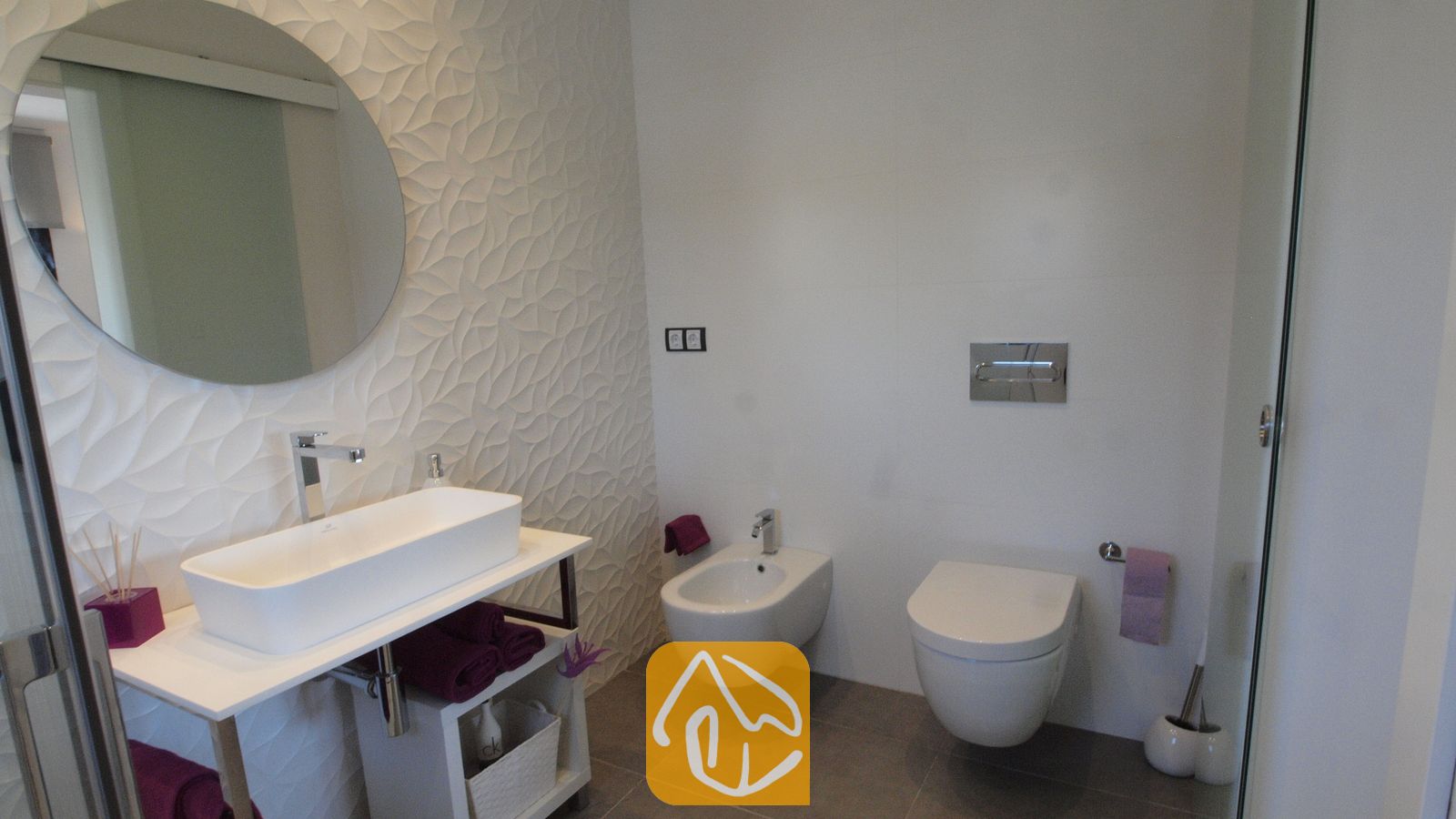 Vakantievilla's Costa Brava Spanje - Villa SummerDream - Moderne badkamer met een witte opzetwastafel op een wastafelmeubel, een ronde spiegel, een bidet en een wandcloset tegen een achtergrond van getextureerde witte tegels.