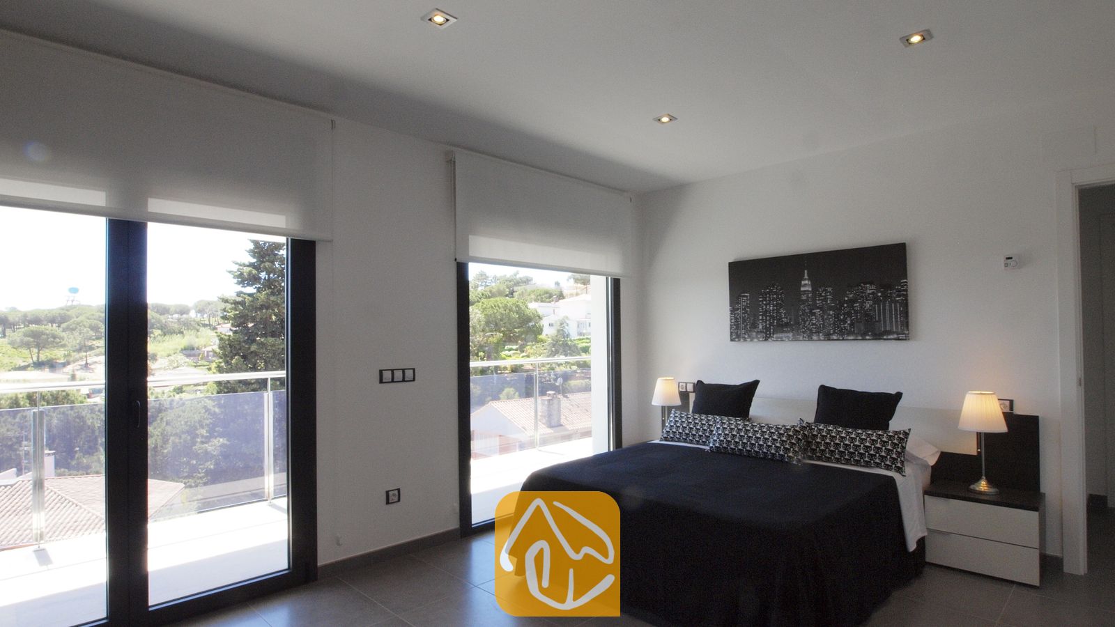 Vakantievilla's Costa Brava Spanje - Villa SummerDream - Slaapkamer met een tweepersoonsbed, zwart-witte beddengoed, bijpassende nachtkastjes met lampen en grote glazen deuren die uitkomen op een balkon met uitzicht op groen.
