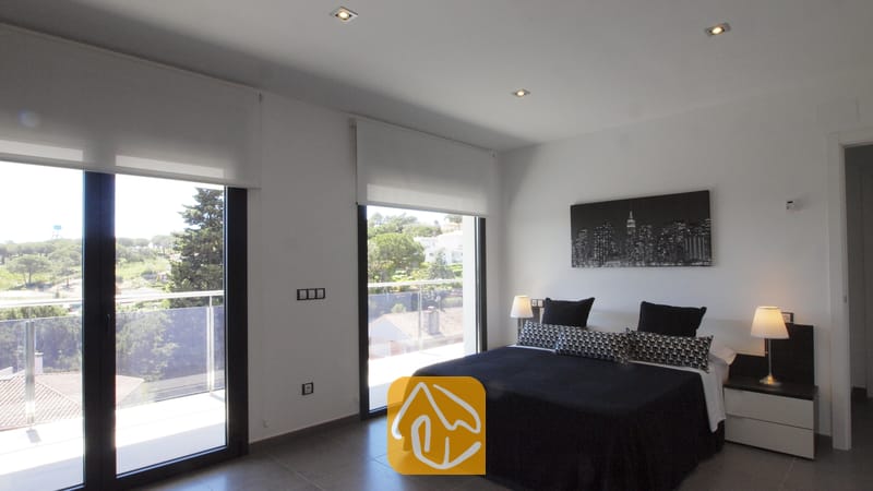 Villas de vacances Costa Brava Espagne - Villa SummerDream - Chambre avec un lit double, du linge de lit noir et blanc, des tables de chevet assorties avec lampes, et de grandes portes vitrées s'ouvrant sur un balcon avec vue sur la verdure.