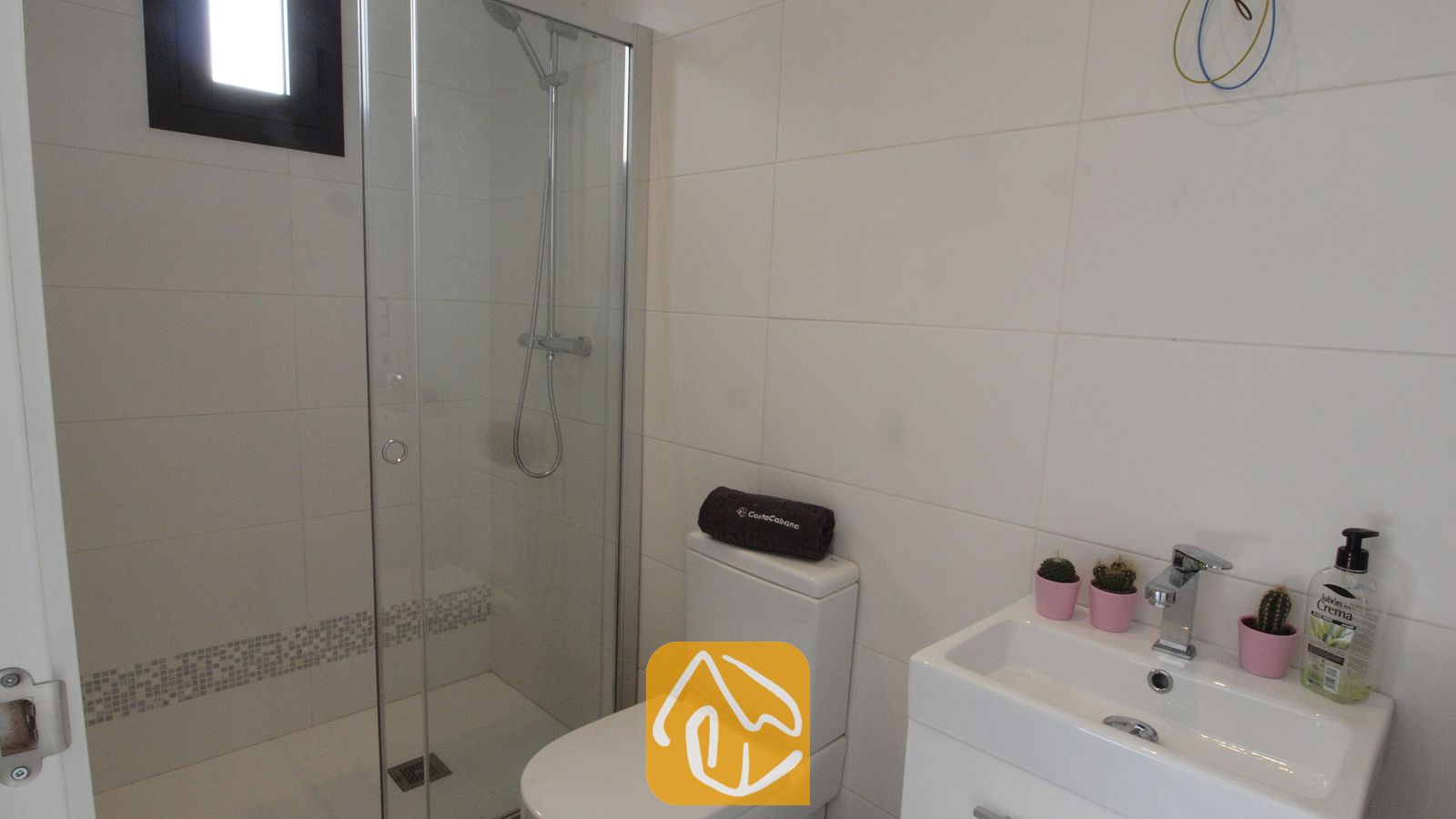 Vakantievilla's Costa Brava Spanje - Villa SummerDream - Moderne badkamer met een inloopdouche met glazen deur, wit getegelde muren, een toilet met een opgerolde donkere handdoek erop, en een witte wastafel met roze cactussen in pot.
