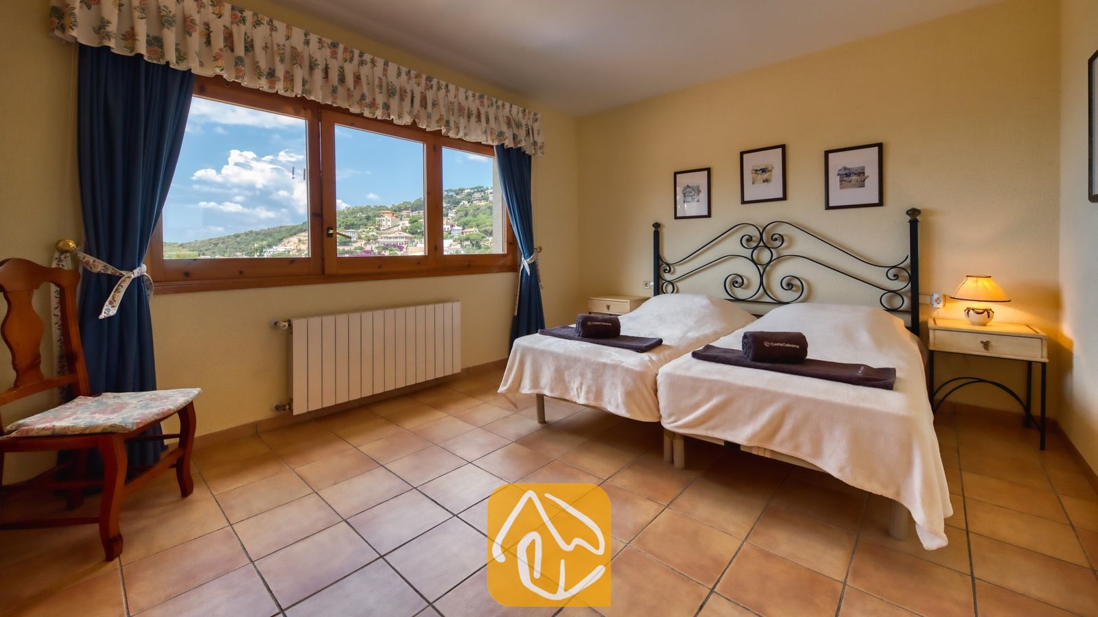 Vakantievilla's Costa Brava Spanje - Villa Cleo - Slaapkamer met twee eenpersoonsbedden voorzien van een zwart metalen hoofdbord, tegelvloeren en een groot raam dat uitzicht biedt op de heuvels.