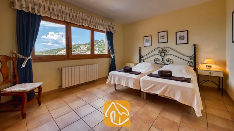 Vakantievilla's Costa Brava Spanje - Villa Cleo - Slaapkamer met twee eenpersoonsbedden voorzien van een zwart metalen hoofdbord, tegelvloeren en een groot raam dat uitzicht biedt op de heuvels.