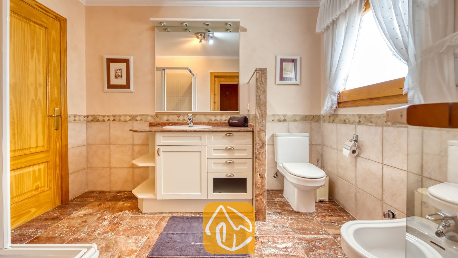 Vakantievilla's Costa Brava Spanje - Villa Cleo - Badkamer voorzien van een wit badkamermeubel met lades, een spiegel aan de wand, beige betegelde muren en bruine vloertegels met een marmerpatroon.