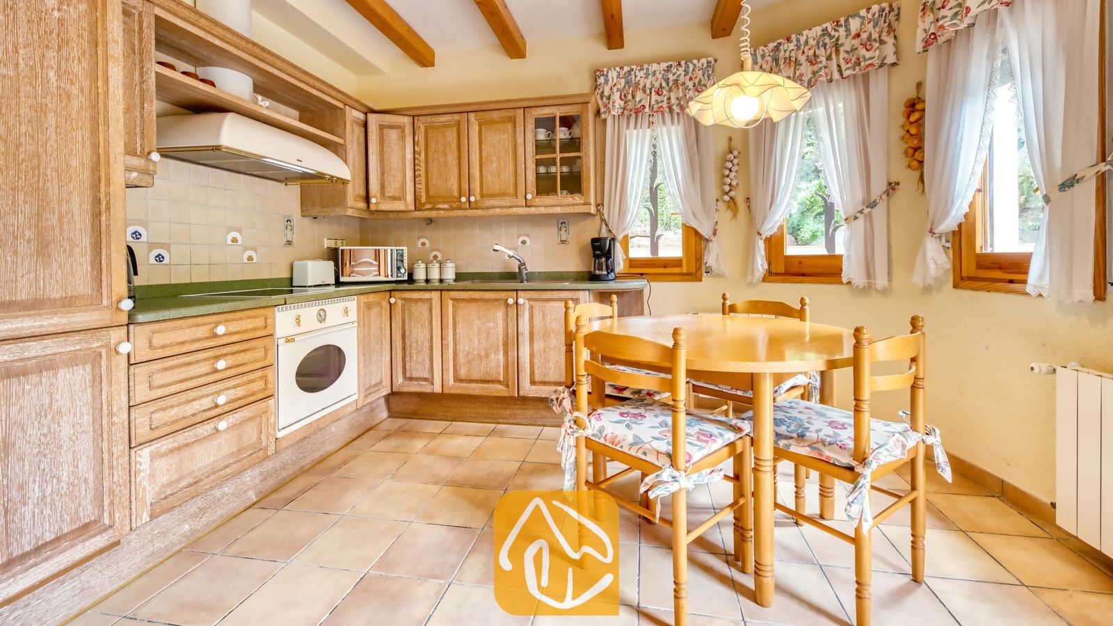 Vakantievilla's Costa Brava Spanje - Villa Cleo - Rustieke keuken met houten kasten, tegelvloeren en een ronde eettafel omringd door vier stoelen met kussens met een bloemenprint.
