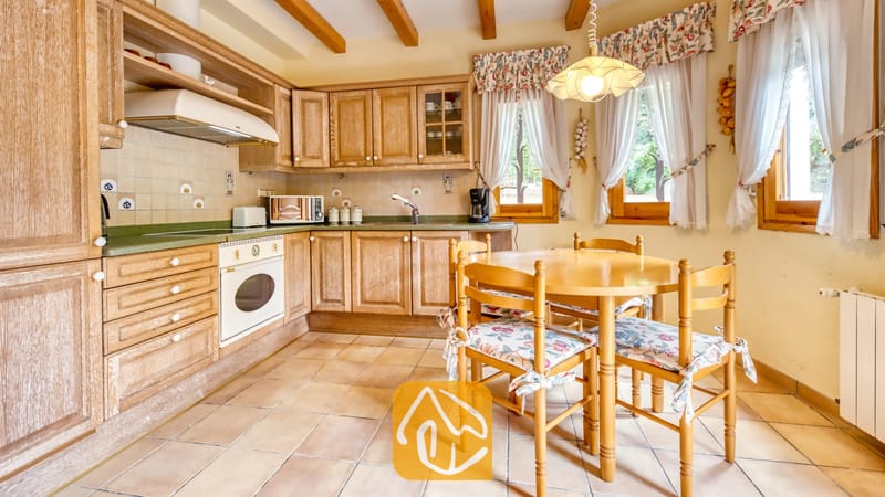 Vakantievilla's Costa Brava Spanje - Villa Cleo - Rustieke keuken met houten kasten, tegelvloeren en een ronde eettafel omringd door vier stoelen met kussens met een bloemenprint.