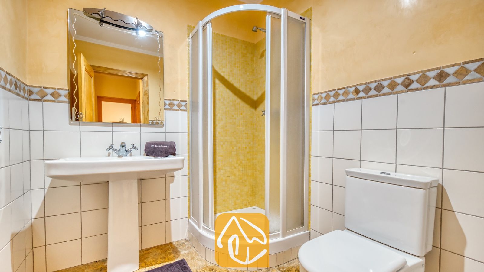 Vakantievilla's Costa Brava Spanje - Villa Cleo - Badkamer met een hoekdouche met gele mozaïektegels, een witte wastafel op zuil met daarboven een spiegel, en een wit toilet tegen tegelmuren.