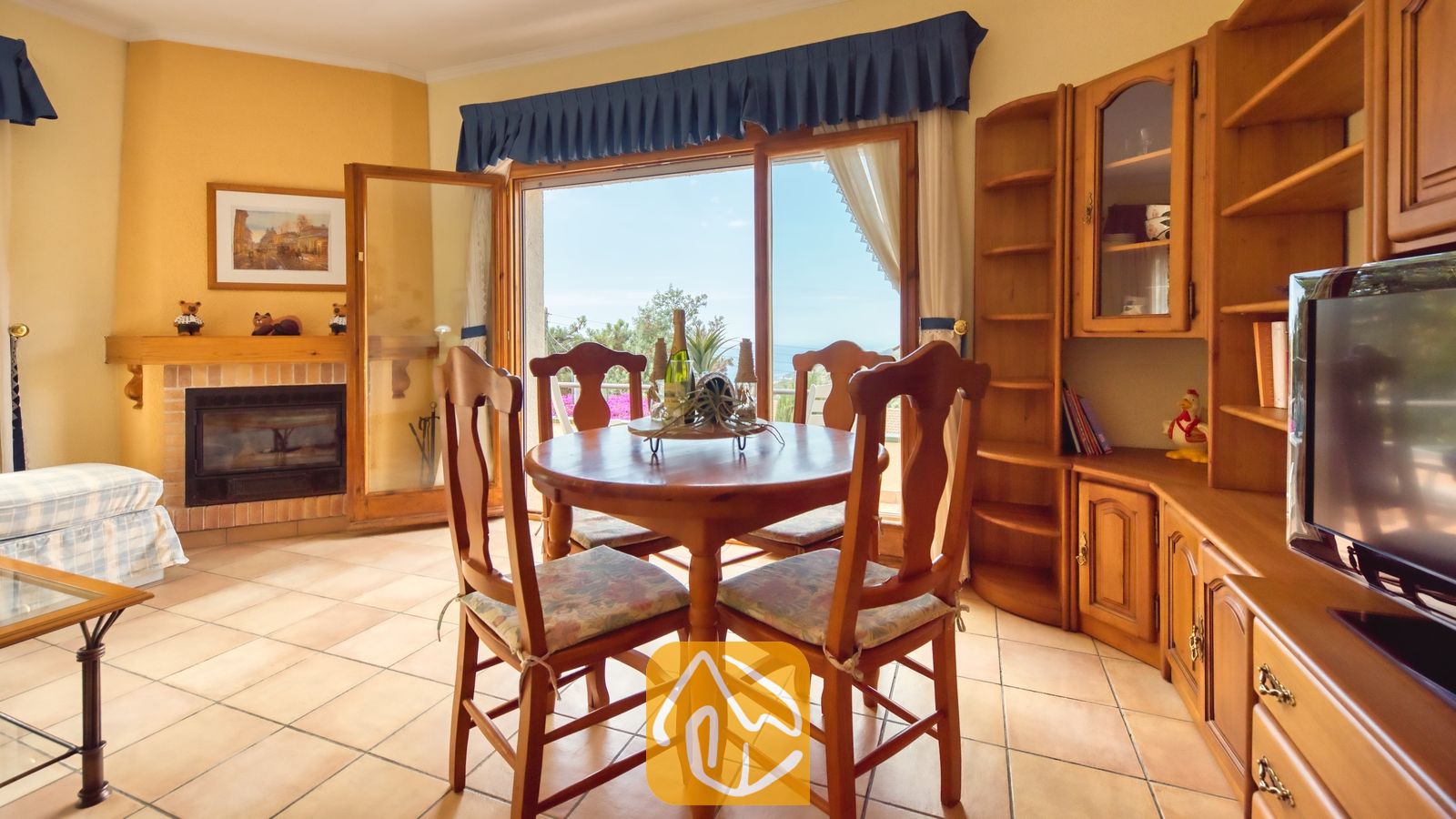 Vakantievilla's Costa Brava Spanje - Villa Cleo - Ronde houten eettafel met vier bijpassende stoelen in Villa Cleo, voorzien van een stenen open haard en grote glazen schuifdeuren die naar buiten openen.