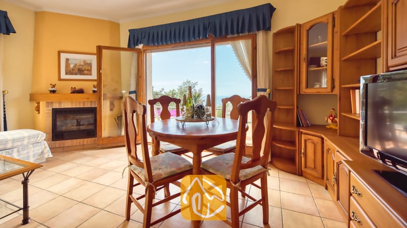 Vakantievilla's Costa Brava Spanje - Villa Cleo - Ronde houten eettafel met vier bijpassende stoelen in Villa Cleo, voorzien van een stenen open haard en grote glazen schuifdeuren die naar buiten openen.