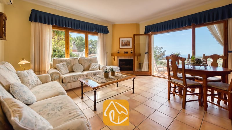 Vakantievilla's Costa Brava Spanje - Villa Cleo - Woon- en eetkamer in Villa Cleo, voorzien van tegelvloeren, banken met een bloemendessin, een centrale glazen salontafel, een houten eettafel en grote ramen met uitzicht op buiten.