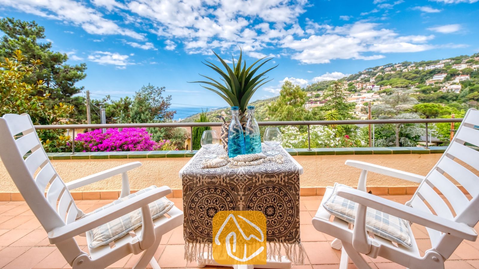 Vakantievilla's Costa Brava Spanje - Villa Cleo - Balkonterras bij Villa Cleo met een klein tafeltje met een tafelkleed met motief, twee witte stoelen met kussens en uitzicht op zee op de achtergrond.