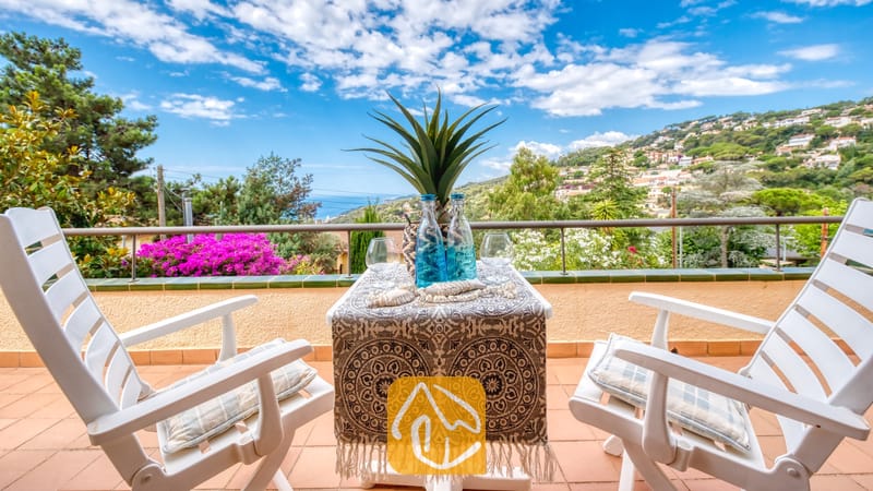 Vakantievilla's Costa Brava Spanje - Villa Cleo - Balkonterras bij Villa Cleo met een klein tafeltje met een tafelkleed met motief, twee witte stoelen met kussens en uitzicht op zee op de achtergrond.