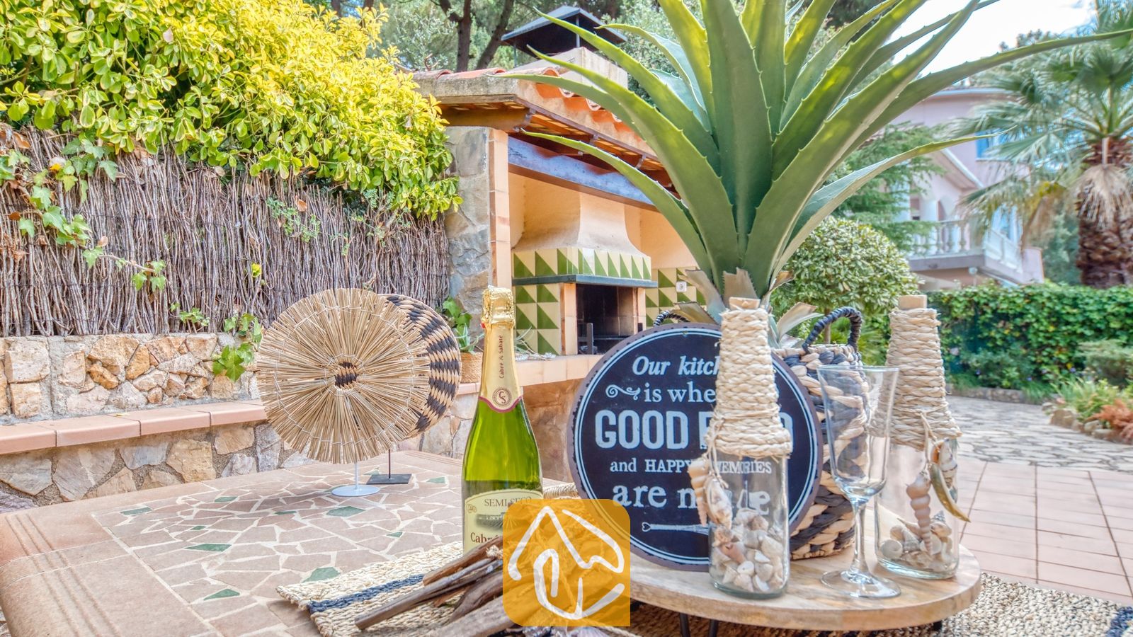 Vakantievilla's Costa Brava Spanje - Villa Cleo - Buitentuinserettafel met een fles champagne, een decoratief bord, glazen flessen gevuld met schelpen en een stenen barbecue op de achtergrond bij Villa Cleo.