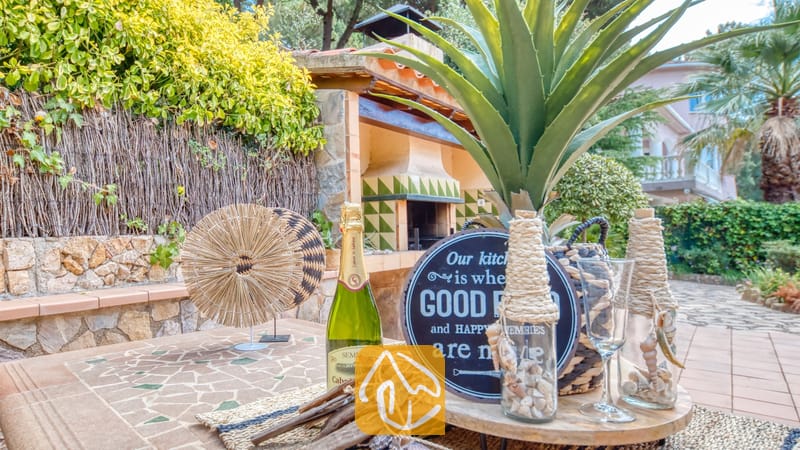 Vakantievilla's Costa Brava Spanje - Villa Cleo - Buitentuinserettafel met een fles champagne, een decoratief bord, glazen flessen gevuld met schelpen en een stenen barbecue op de achtergrond bij Villa Cleo.