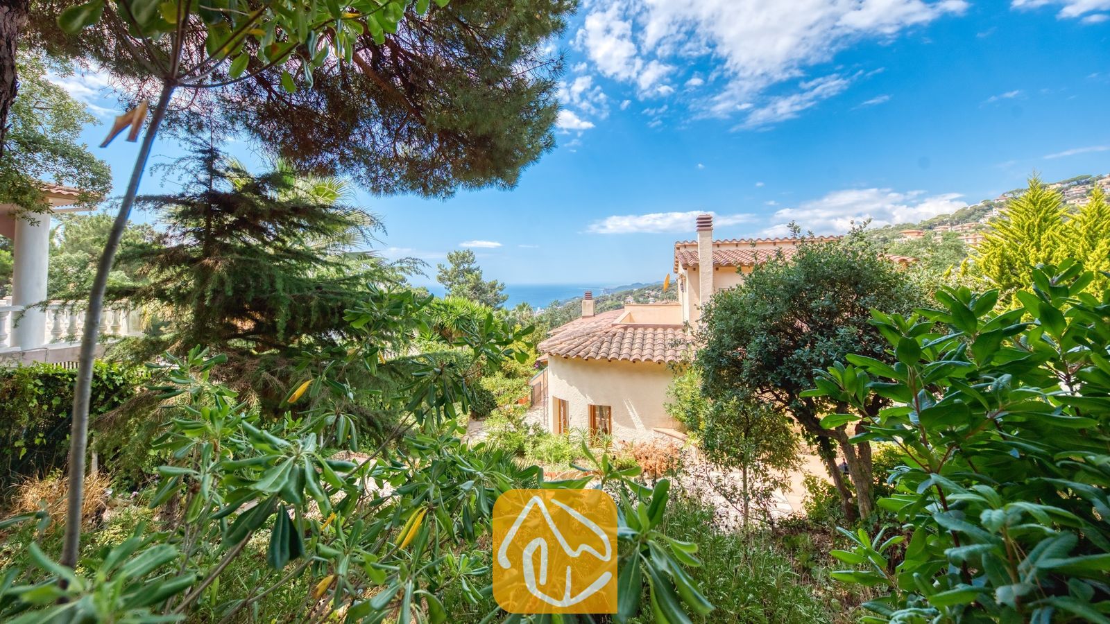 Vakantievilla's Costa Brava Spanje - Villa Cleo - Buitenaanzicht van Villa Cleo in Spanje, omlijst door weelderig groen tuingroen met traditionele terracotta dakpannen en de zee zichtbaar op de achtergrond onder een helderblauwe hemel.